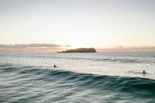 Eine Weitwinkelaufnahme des Ozeans bei Sonnenuntergang. Zwei Surfer auf Brettern sind im Wasser, mit einer großen Welle im