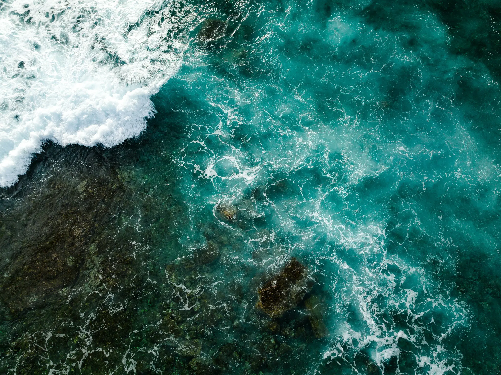 An aerial shot shows a large white ocean wave crashing over dark, submerged rocks, with clear turquoise water beyond.