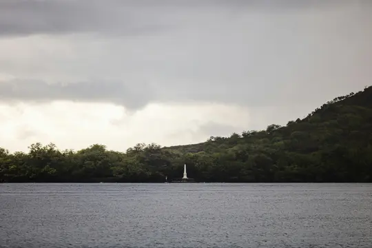 Ein weißes Obelisk-Denkmal steht prominent auf einer kleinen, baumbestandenen Landmasse, mit dunkelblauem Wasser im