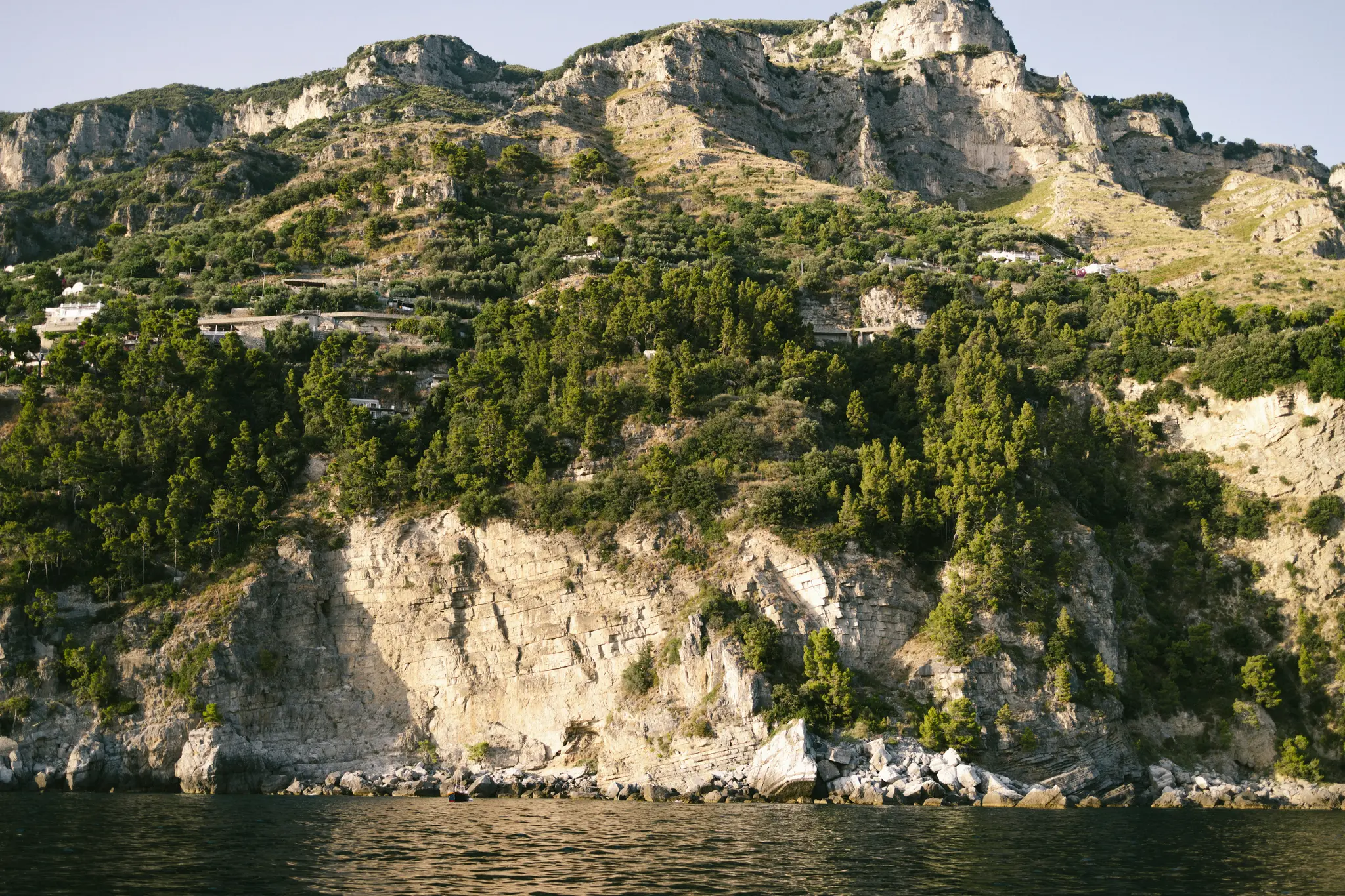 En bred landskapsvy över ett kargt, trädbevuxet berg som reser sig brant ur det mörkblå havet under en klar himmel.