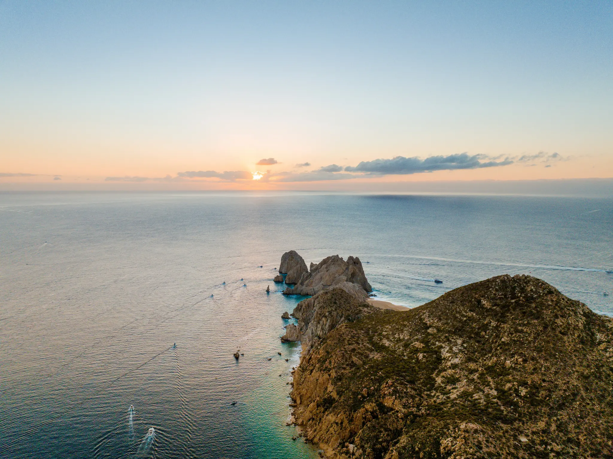 Una foto aerea mostra un tramonto su un vasto oceano, con una costa rocciosa e frastagliata e una piccola spiaggia sabbiosa