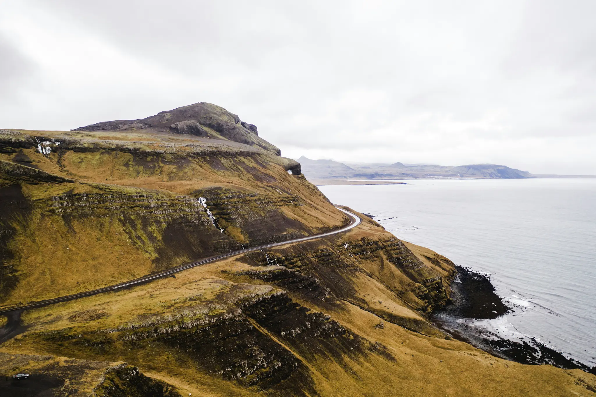 An aerial view shows a dark, winding road cutting across a steep, grassy hillside that meets a grey ocean. The sky is…