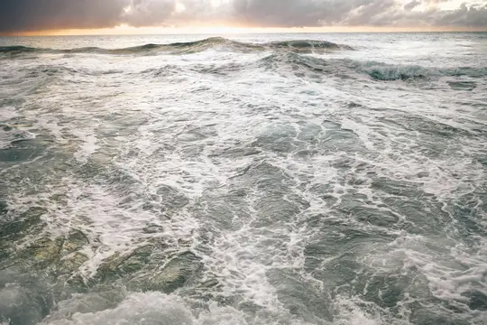 A wide shot of the ocean with numerous waves and white foam covering the surface. The sky above is cloudy with hints of…