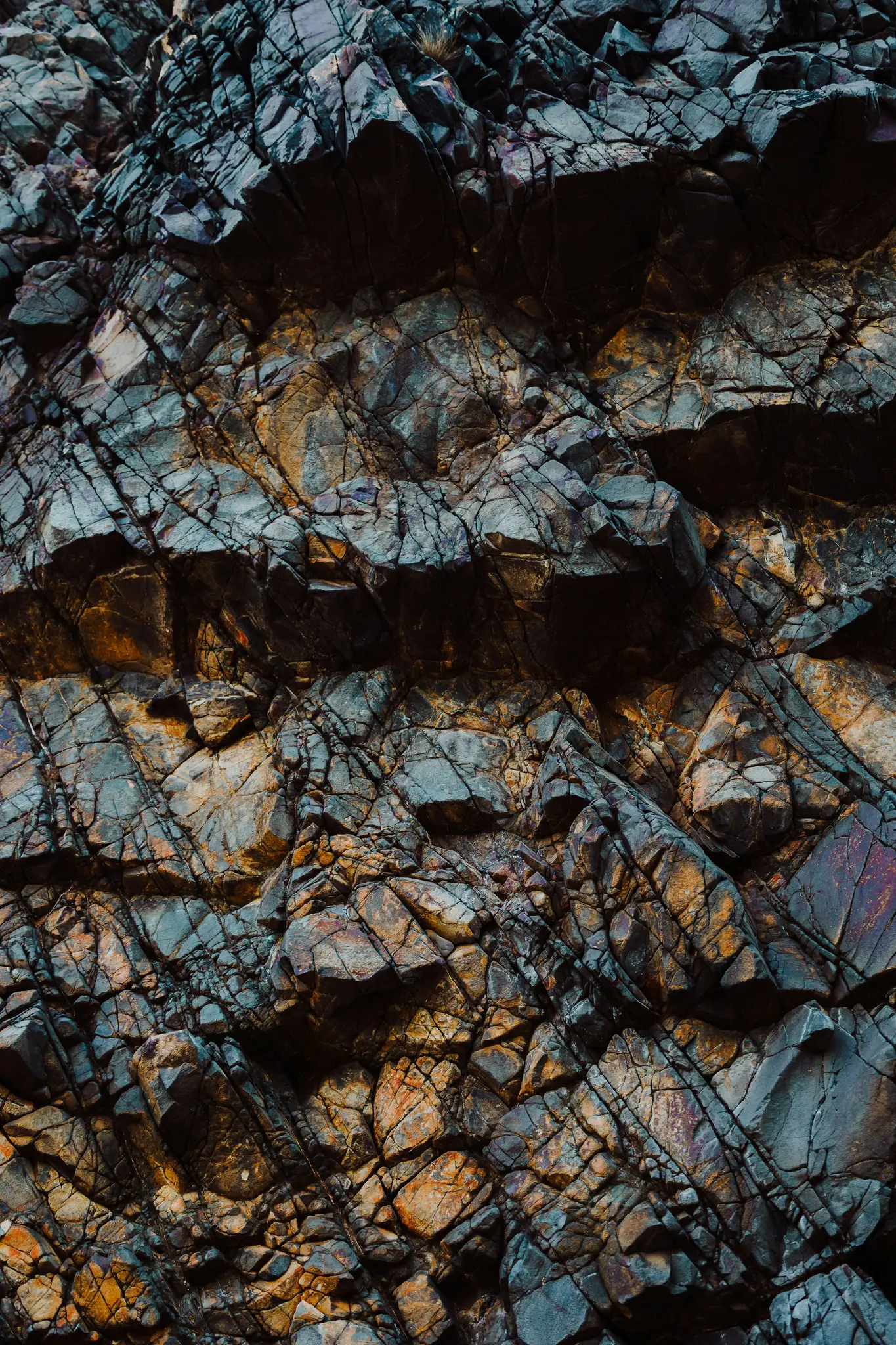 A detailed close-up of a rock face, showing a complex pattern of cracks and fissures in dark blue, gray, and orange tones.