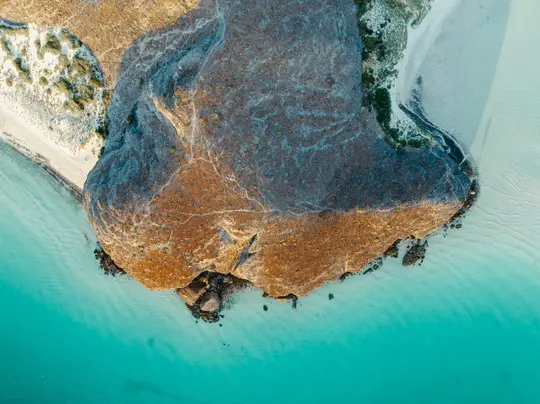 Eine Luftaufnahme zeigt ein großes, braunes, felsiges Vorgebirge, das in lebendiges türkisfarbenes Wasser ragt und von hellen