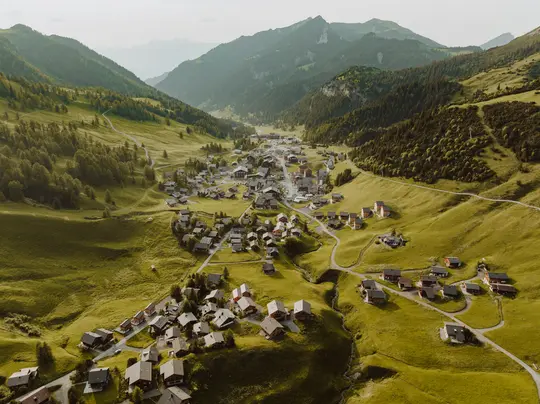 Una vista aerea mostra un piccolo villaggio con molte case sparse in una lussureggiante valle verde, circondata da montagne