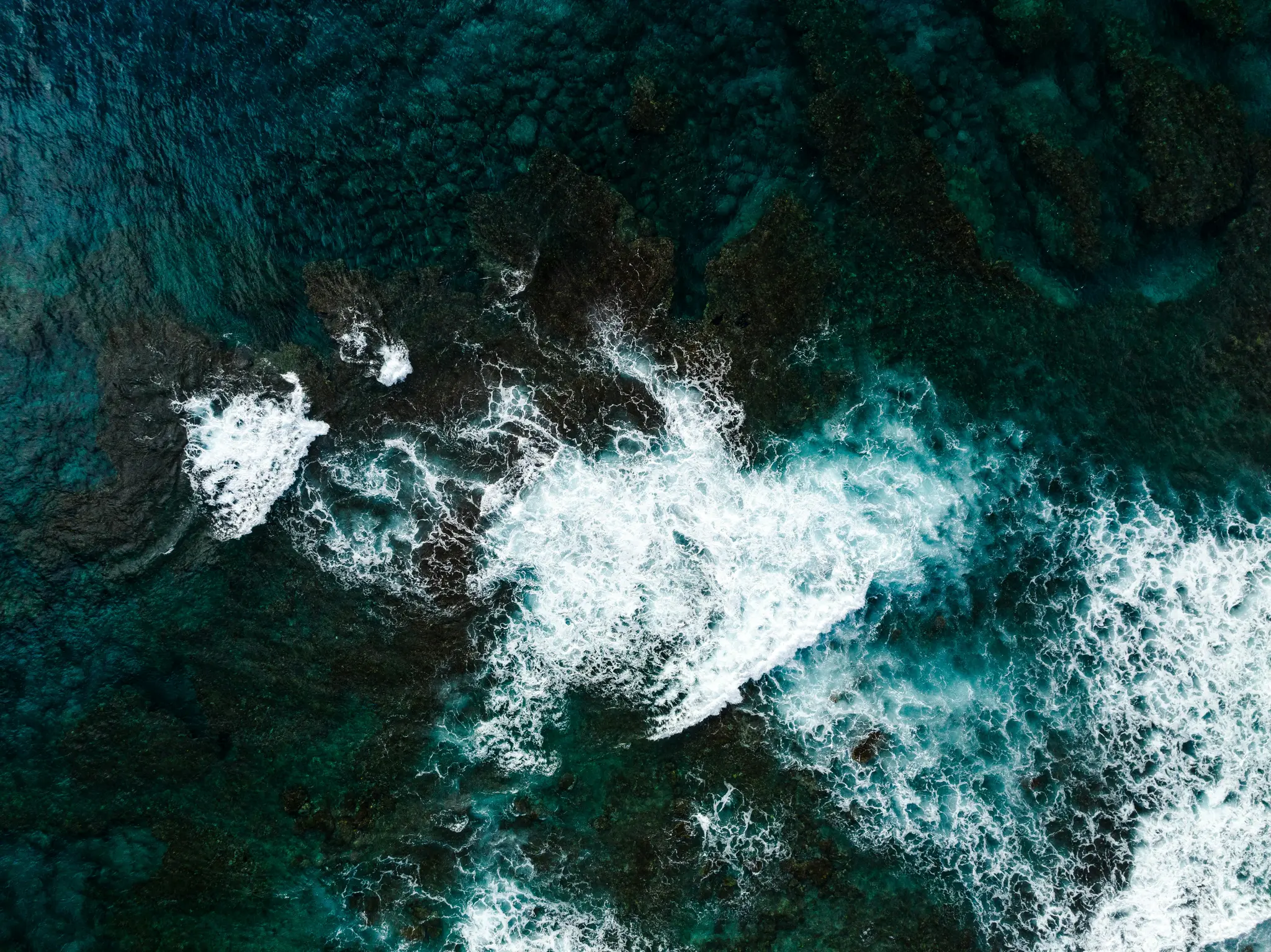 Eine Luftaufnahme zeigt dunkelblaues und grünes Meerwasser mit weißen Wellen, die über dunkle, untergetauchte Felsen und