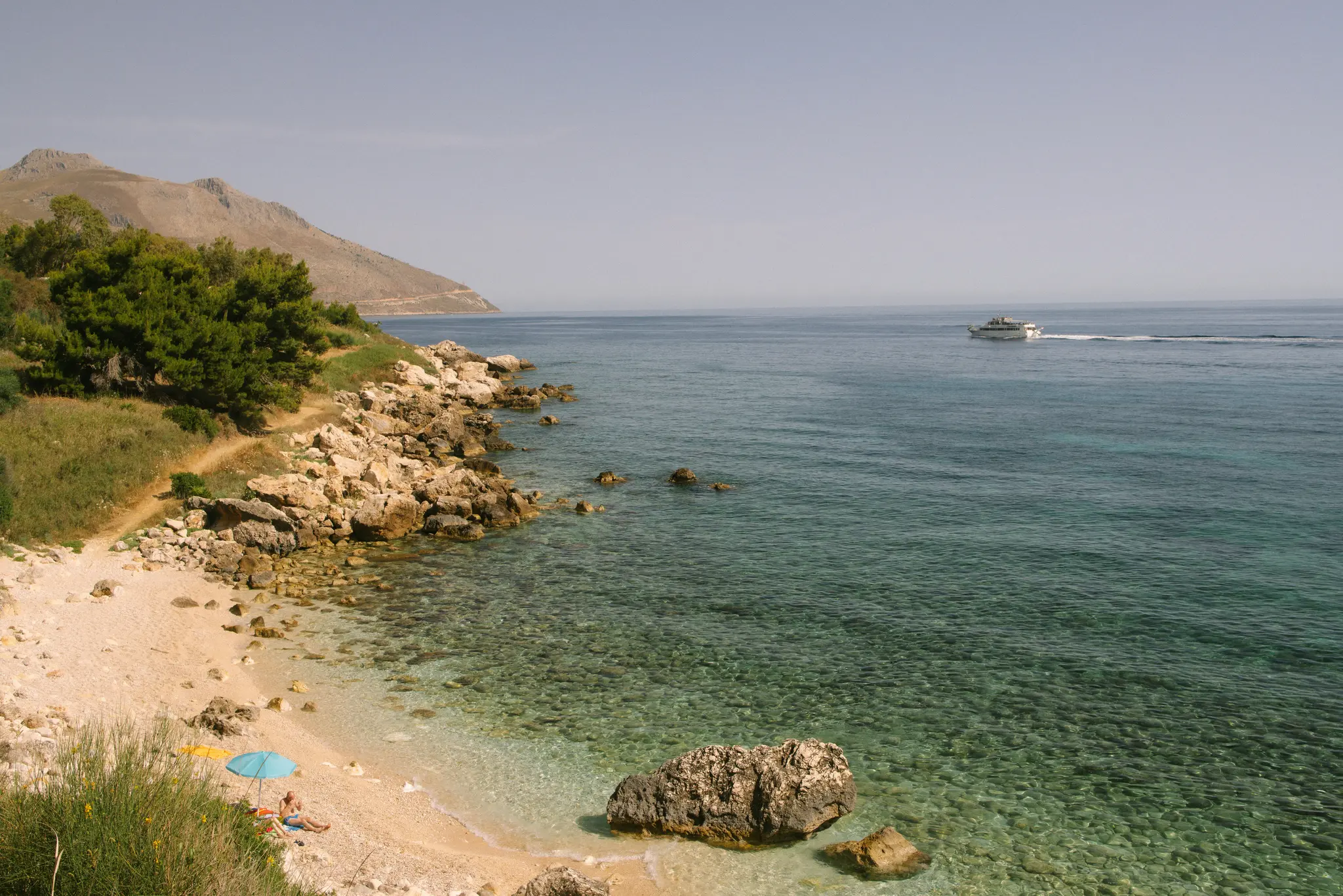 A high-angle view of a rocky coastline with a small pebble beach and clear turquoise water. A person is under a blue…
