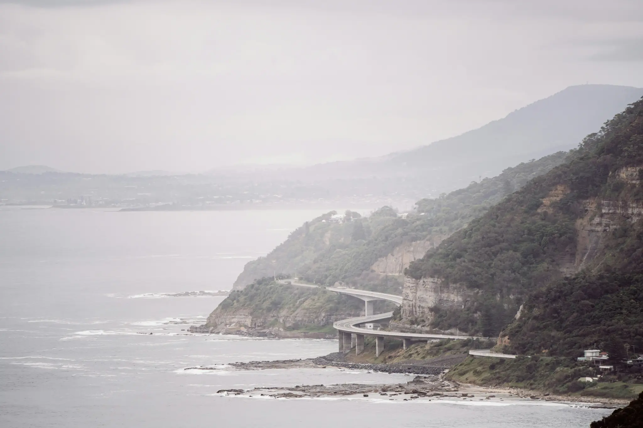 A wide landscape view of a multi-level highway curving along a steep, tree-covered cliffside next to a grey ocean under an…
