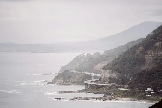 A wide landscape view of a multi-level highway curving along a steep, tree-covered cliffside next to a grey ocean under an…