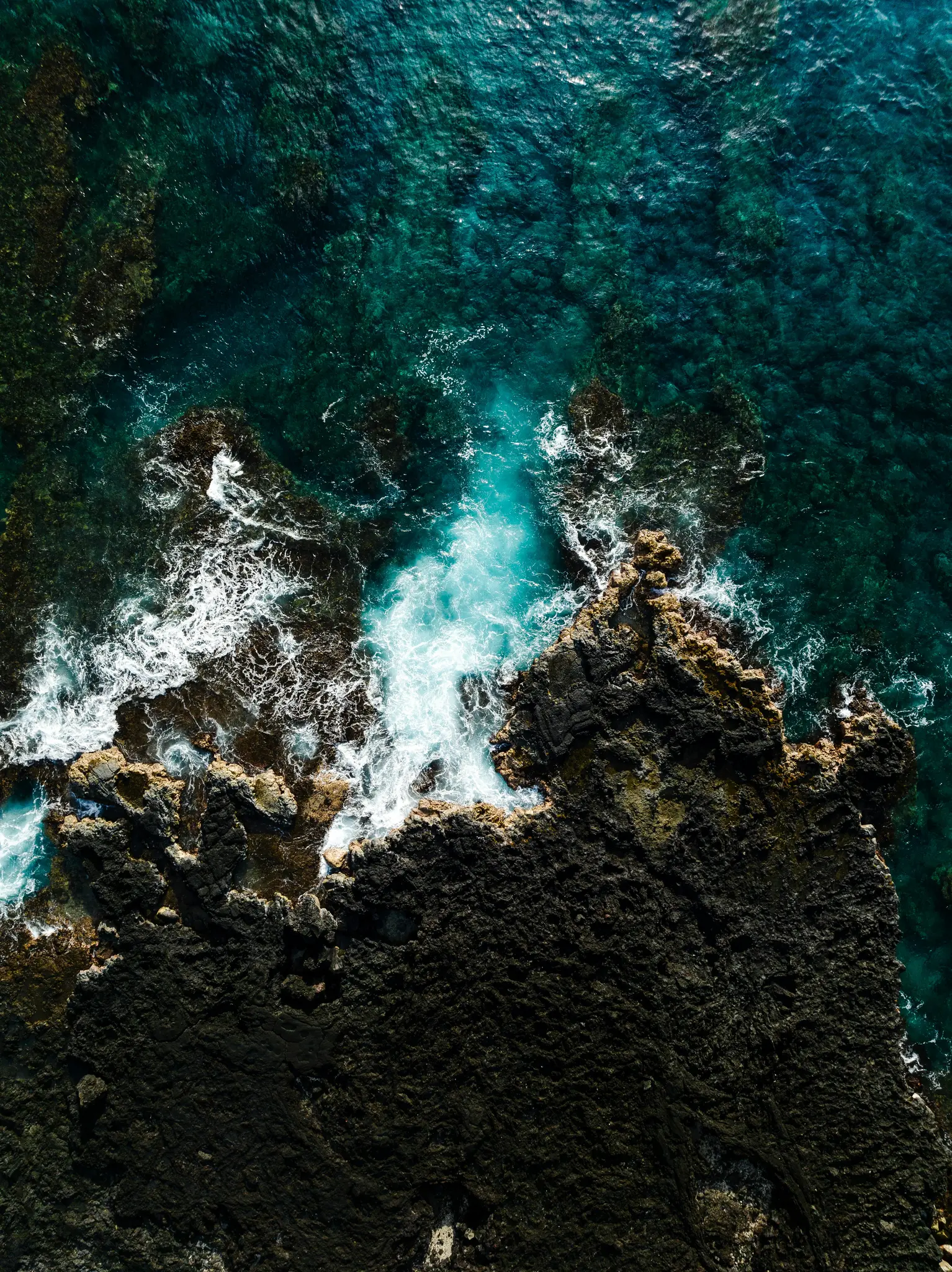 An aerial, top-down view of a rugged volcanic coastline where dark brown and black rocks meet the vibrant blue ocean. White…