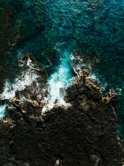 An aerial, top-down view of a rugged volcanic coastline where dark brown and black rocks meet the vibrant blue ocean. White…