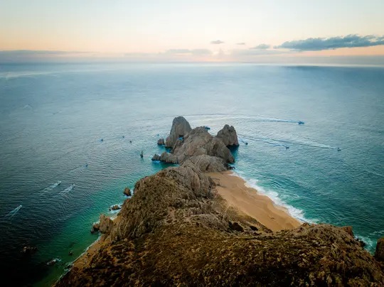 An aerial view shows a rugged brown and tan coastline meeting a sandy beach and turquoise ocean. Rock formations rise from…