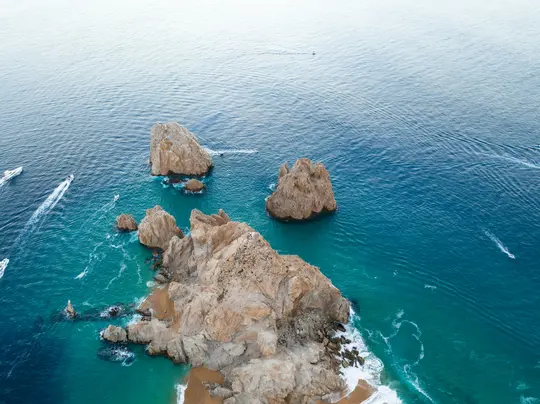 An aerial shot shows a rugged coastline with light brown rock formations jutting out of the turquoise and deep blue ocean.…