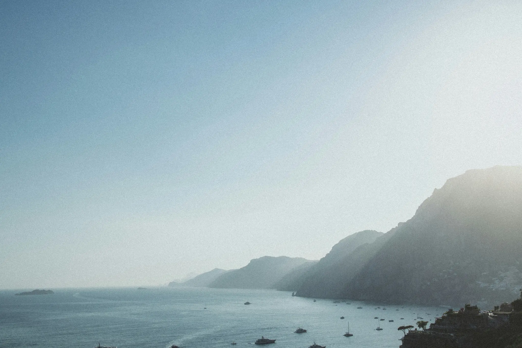 A wide, hazy landscape photo showing a long coastline with dark mountains on the right and a bay with many boats.