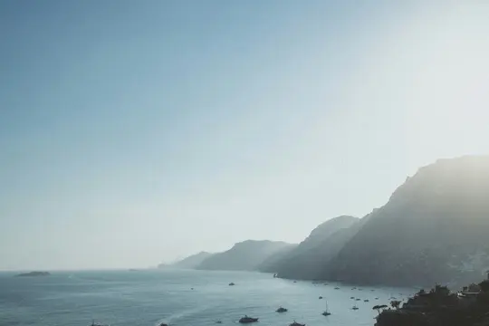 A wide, hazy landscape photo showing a long coastline with dark mountains on the right and a bay with many boats.