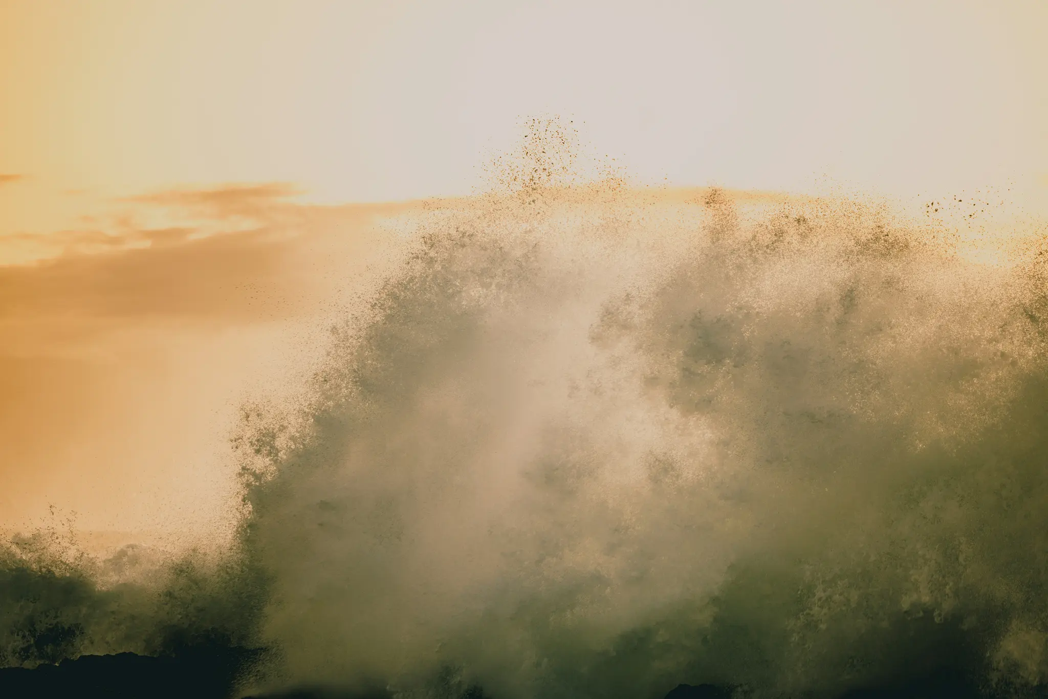 A large ocean wave crashes forcefully, creating a massive spray of water against a warm, orange and yellow sky.