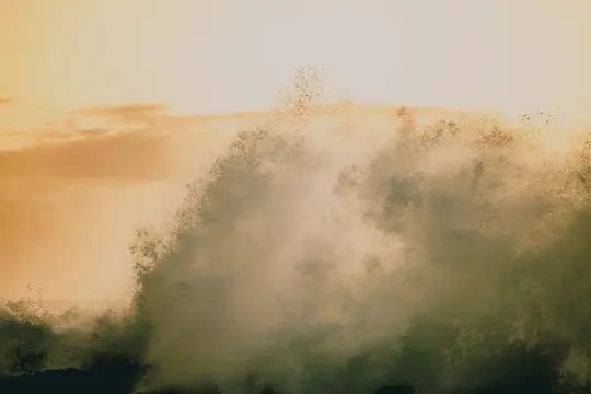 A large ocean wave crashes forcefully, creating a massive spray of water against a warm, orange and yellow sky.