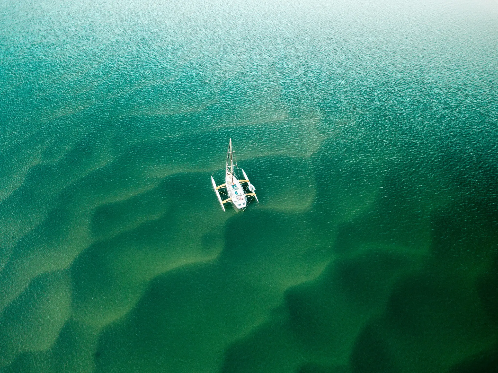 Záber z vtáčej perspektívy na biely trimarán so žltými prvkami plávajúci na čistej, plytkej zelenej vode, odhaľujúci vzory