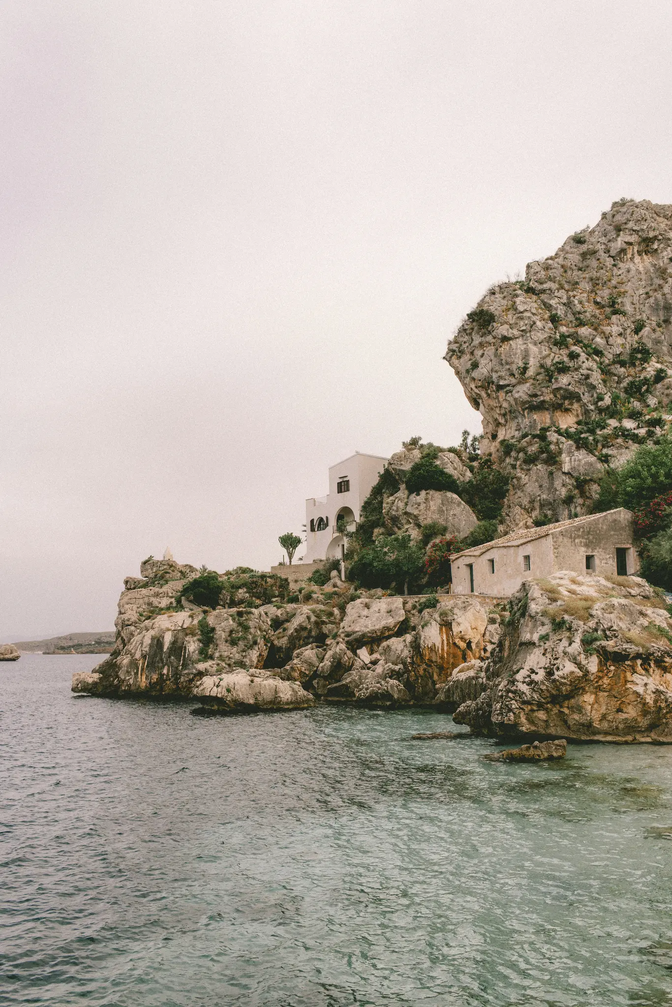 A view of a rocky coastline with two buildings, one white and larger, the other smaller and stone-colored, built into the…