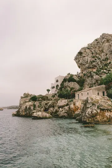 A view of a rocky coastline with two buildings, one white and larger, the other smaller and stone-colored, built into the…