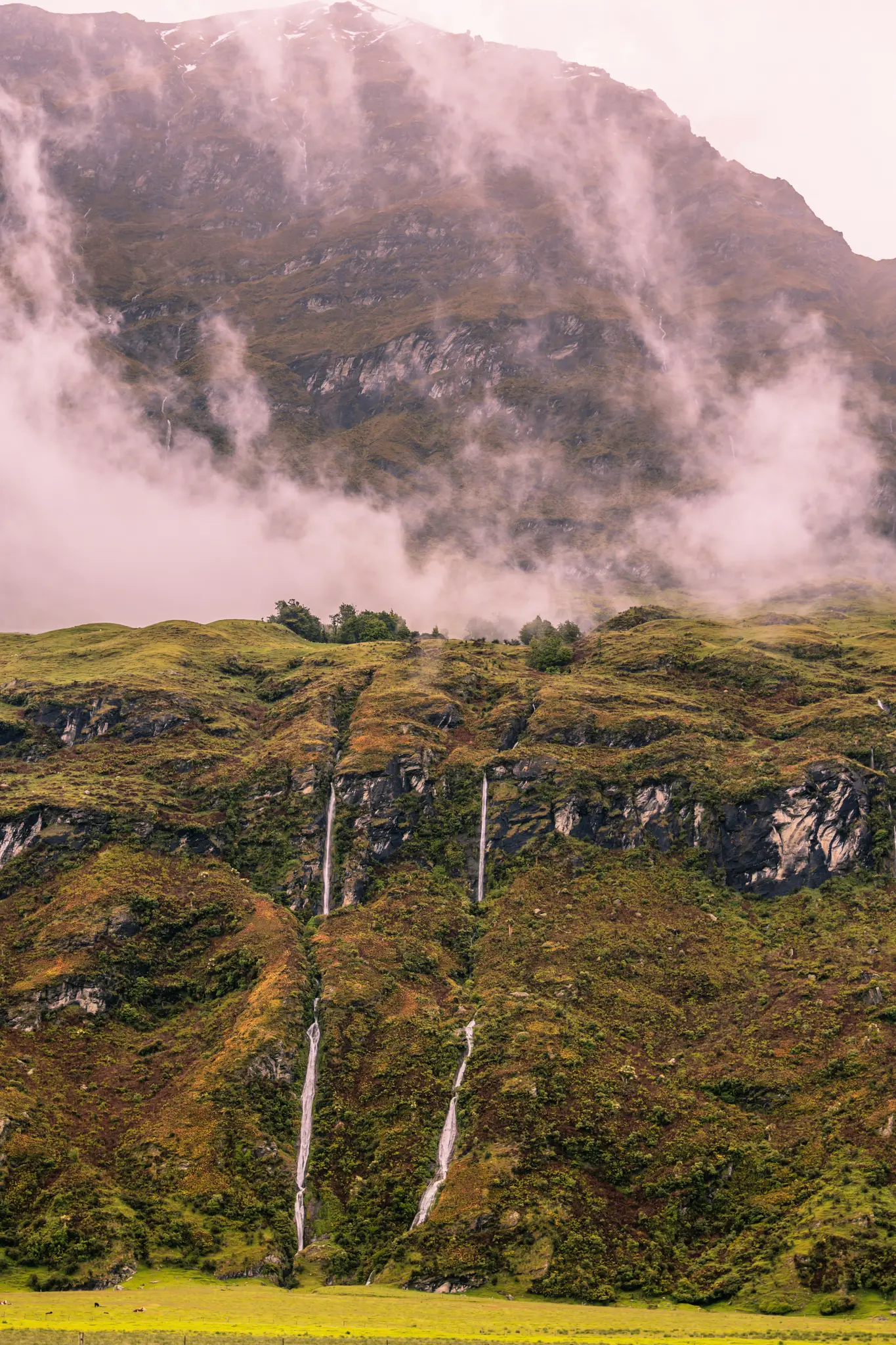 A vertical shot of a lush, green and brown mountain slope with several thin waterfalls cascading down, partially covered by…