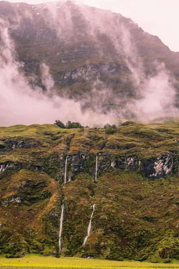 A vertical shot of a lush, green and brown mountain slope with several thin waterfalls cascading down, partially covered by…
