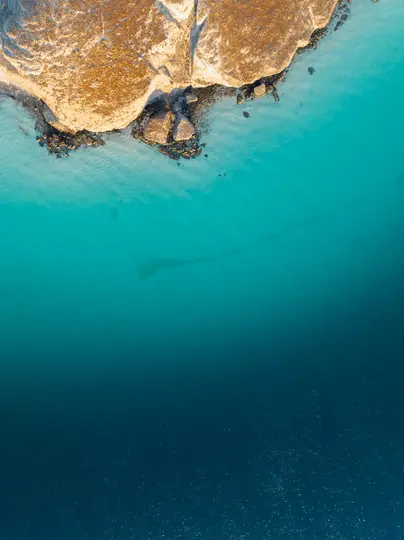 Una ripresa aerea di una costa aspra e marrone chiaro con rocce scure che incontrano acqua turchese limpida che sfuma in un