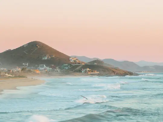 Un'ampia foto di paesaggio di una spiaggia serena con onde dolci, case su colline aride e un cielo morbido rosa e blu.