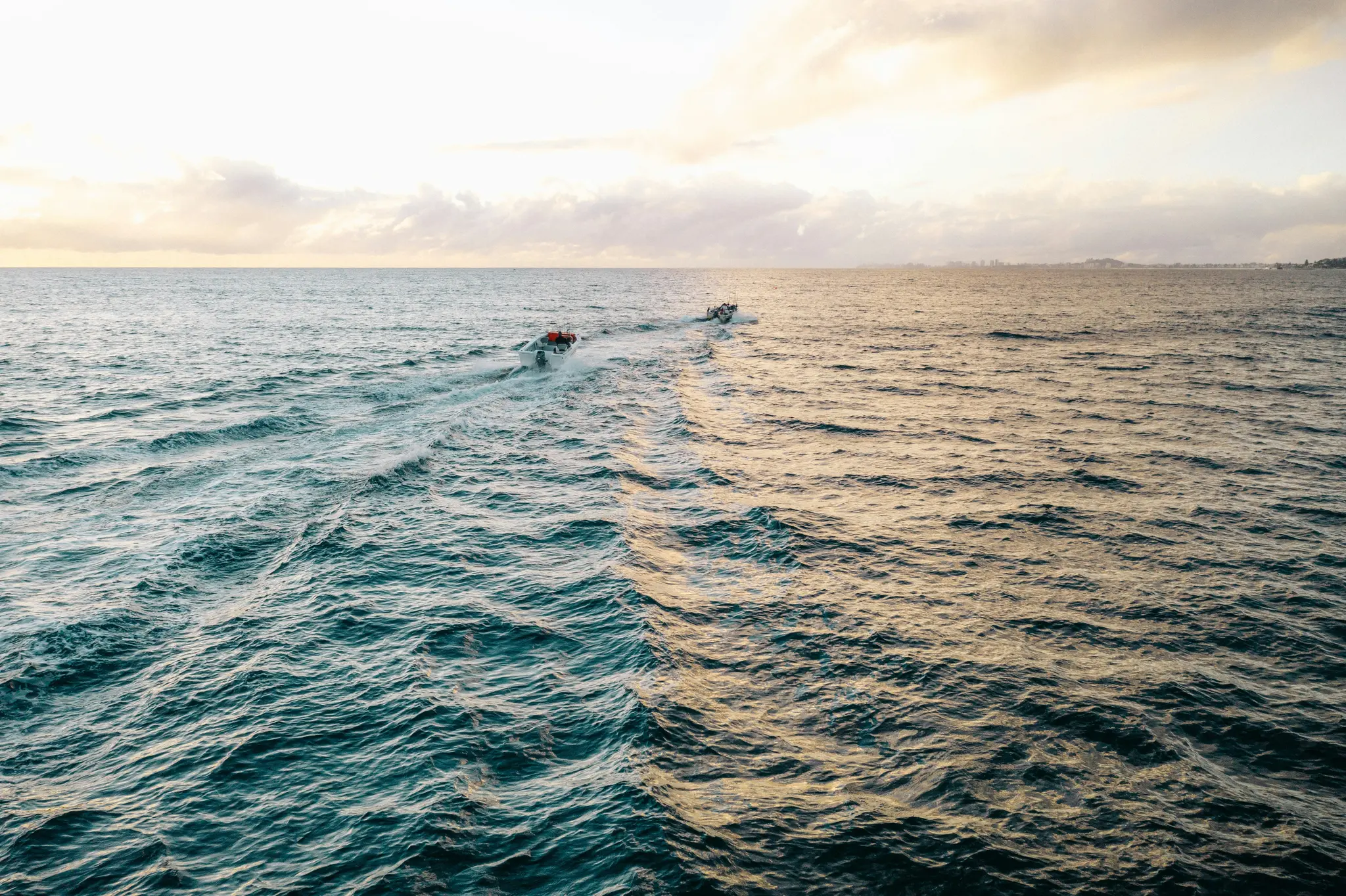 An aerial view of two small boats moving across a vast ocean. The water is teal on the left and golden on the right…