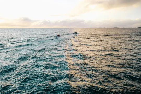 An aerial view of two small boats moving across a vast ocean. The water is teal on the left and golden on the right…