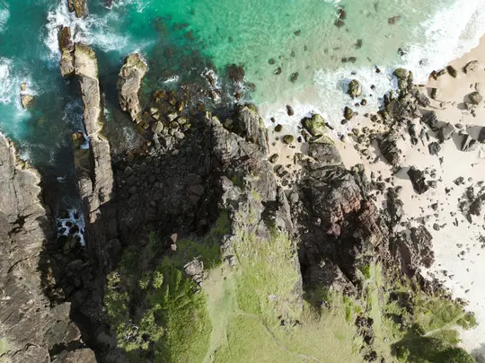 Una vista aerea, dall'alto verso il basso, di una costa frastagliata con rocce scure, vegetazione verde, acqua oceanica