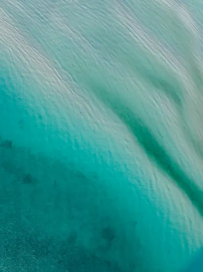 An aerial shot shows clear turquoise ocean water with visible sand ripples on the seabed, transitioning from light to dark…