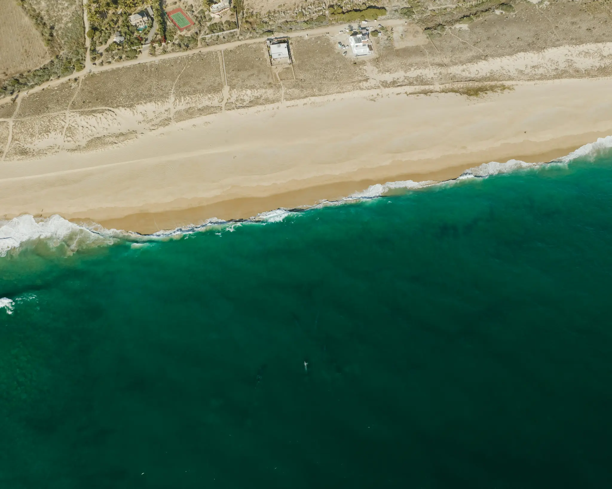 An aerial shot shows a wide beige sandy beach bordering deep teal ocean water with white waves. Inland, dry land with…