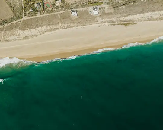 An aerial shot shows a wide beige sandy beach bordering deep teal ocean water with white waves. Inland, dry land with…