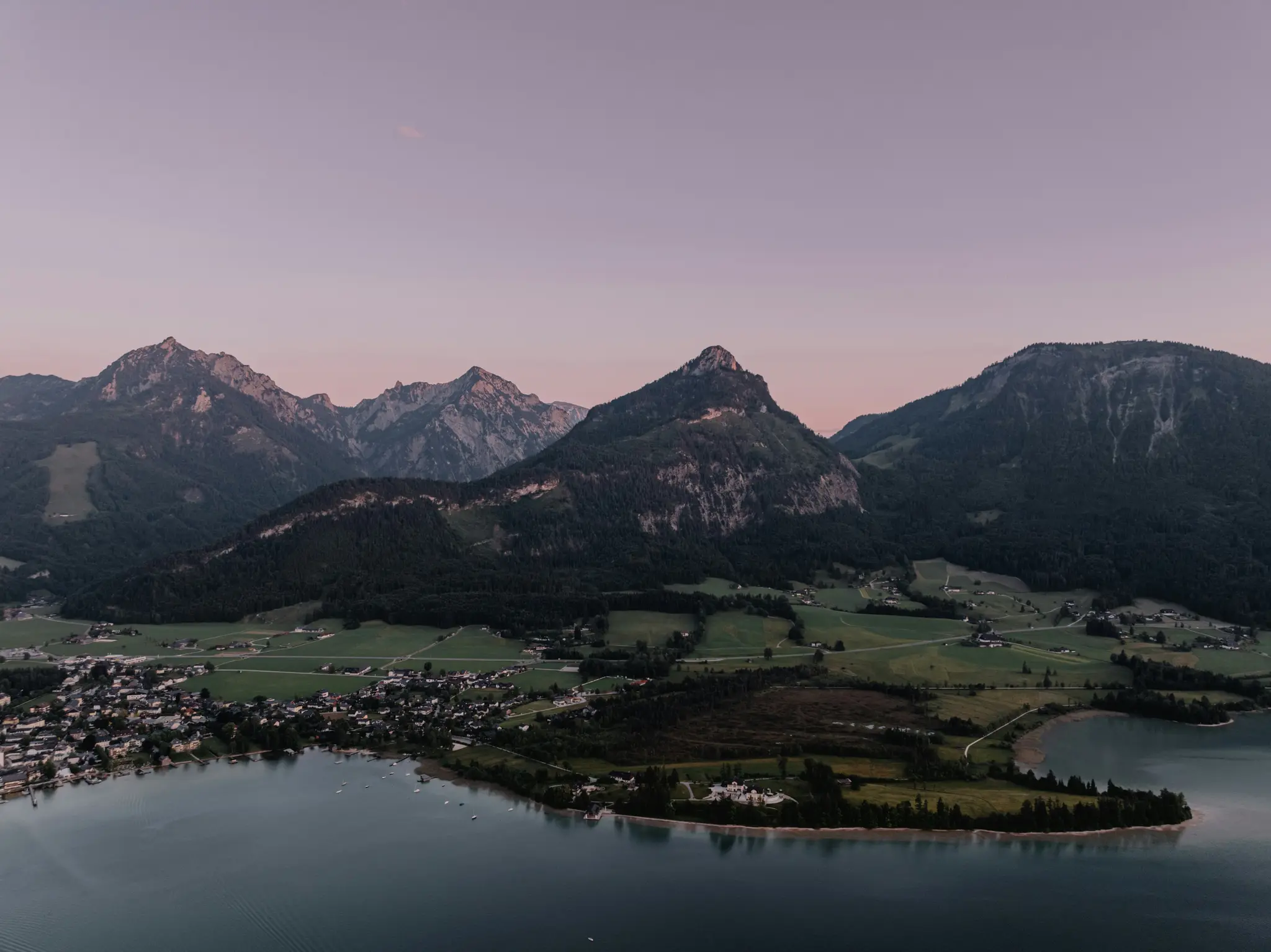An aerial landscape photo shows a dark blue lake with a small town on its left shore, backed by green fields and large…