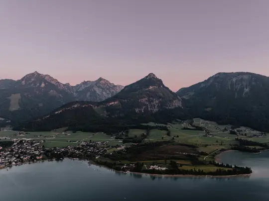 An aerial landscape photo shows a dark blue lake with a small town on its left shore, backed by green fields and large…