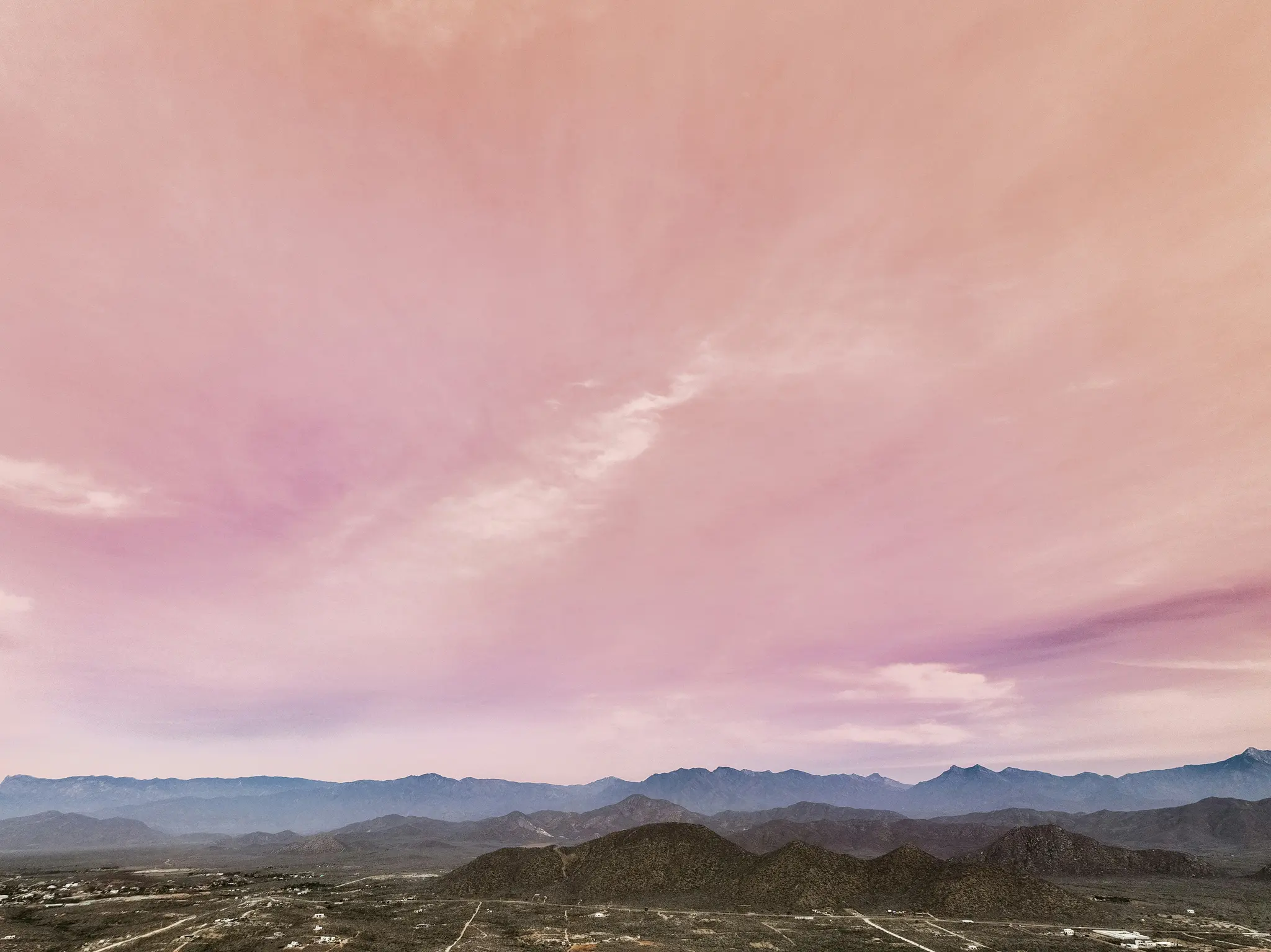 A wide landscape photo showing a vibrant pink and purple sky with wispy clouds over multiple layers of dark blue and green…
