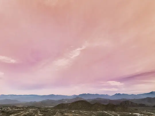 A wide landscape photo showing a vibrant pink and purple sky with wispy clouds over multiple layers of dark blue and green…