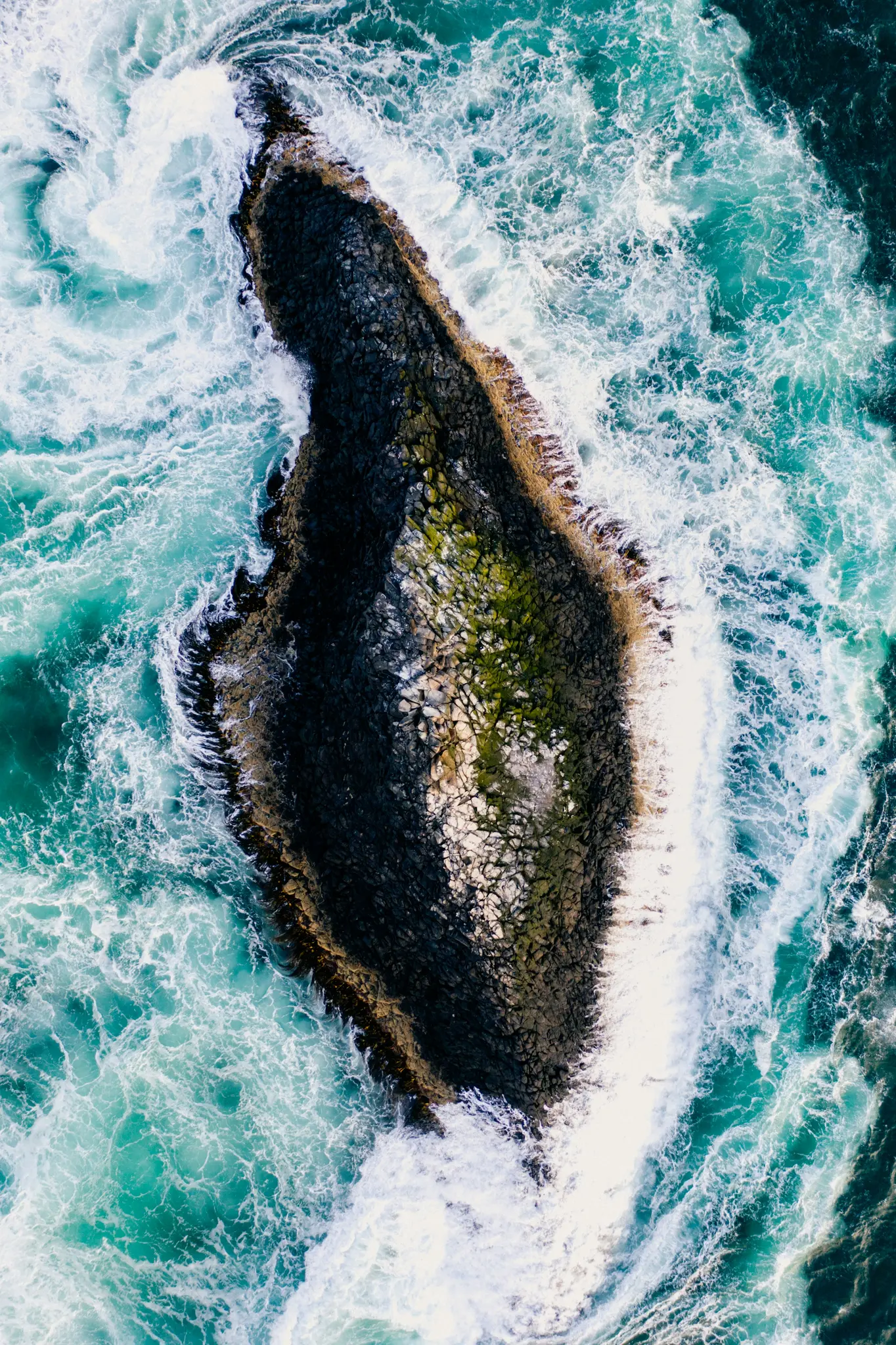 An aerial, top-down view of a dark, elongated rocky island with some green moss, surrounded by swirling turquoise ocean…