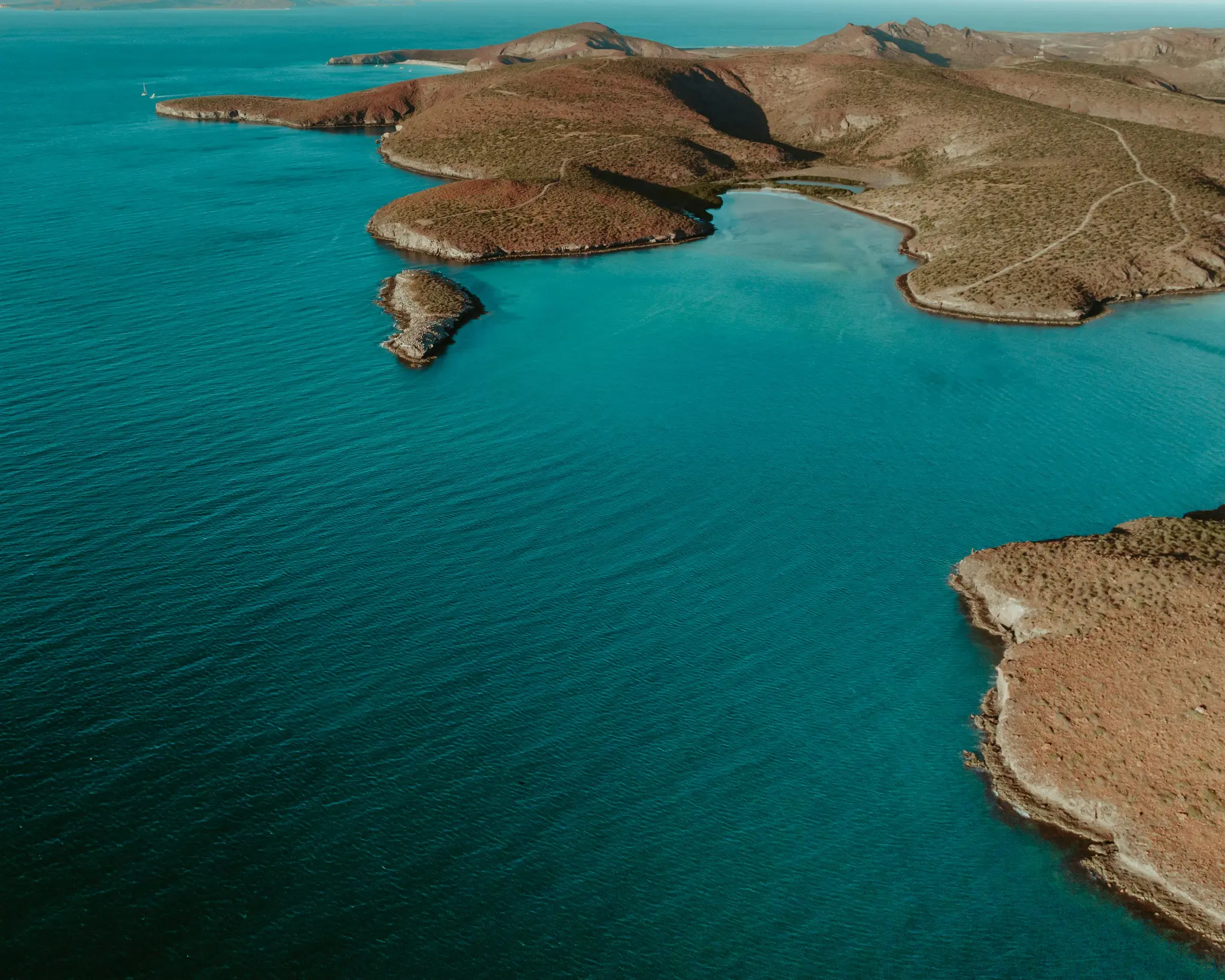 An aerial photograph shows a vibrant turquoise ocean meeting a rugged, arid coastline with reddish-brown hills and sparse…