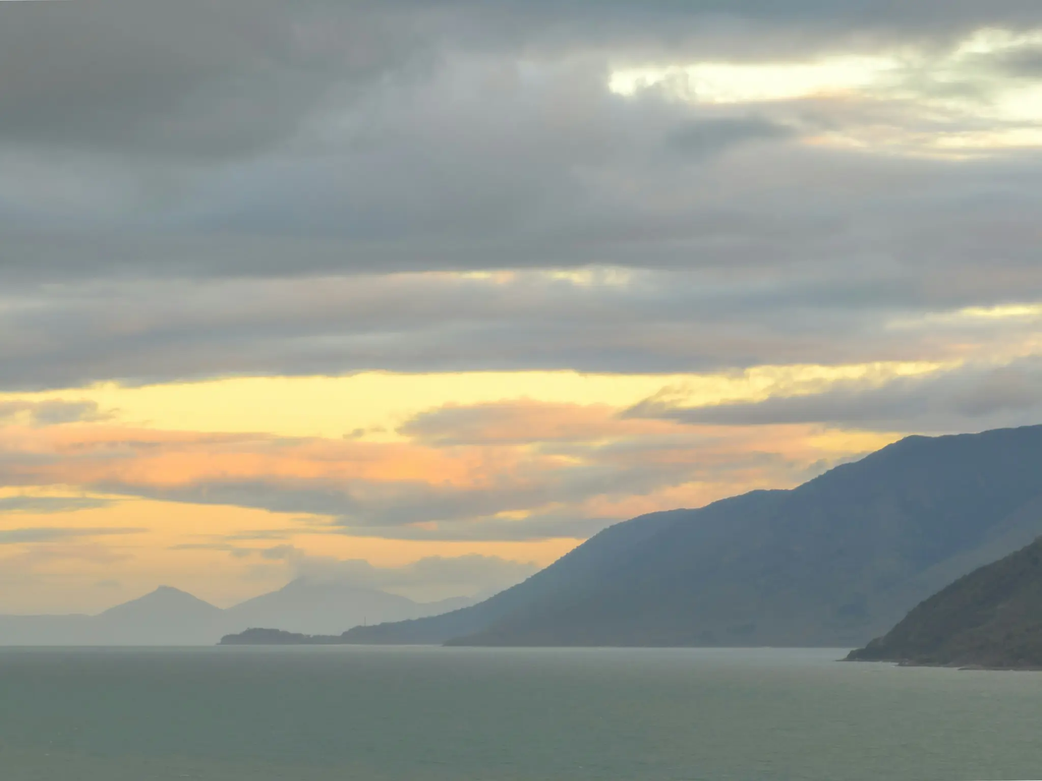 A wide landscape photo showing a calm, greenish-blue sea in the foreground, with a long range of dark blue mountains behind…