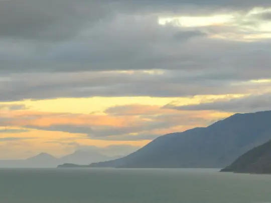 A wide landscape photo showing a calm, greenish-blue sea in the foreground, with a long range of dark blue mountains behind…