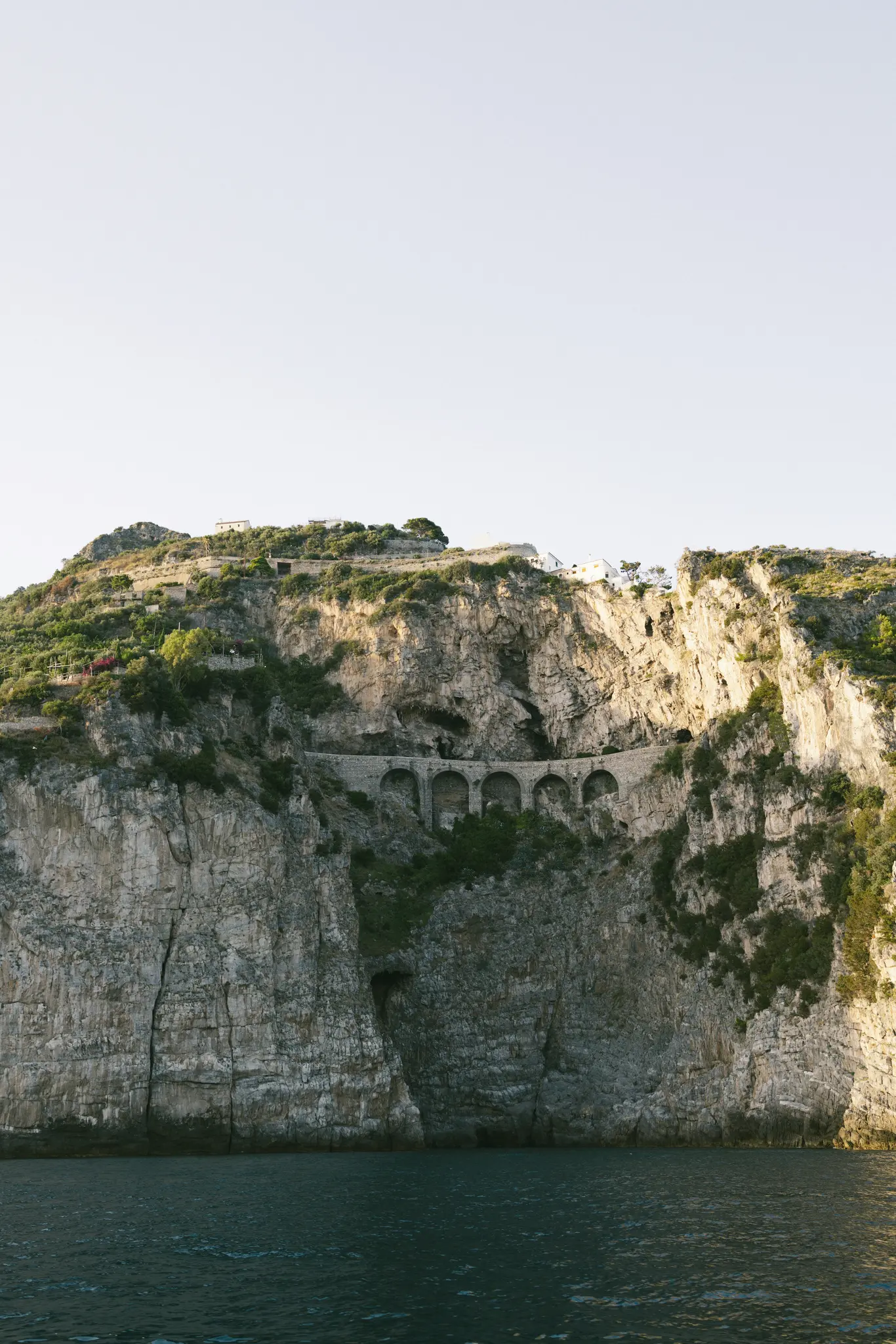A towering, textured cliff face rises from dark blue water under a pale sky. A stone road with multiple arches is carved…