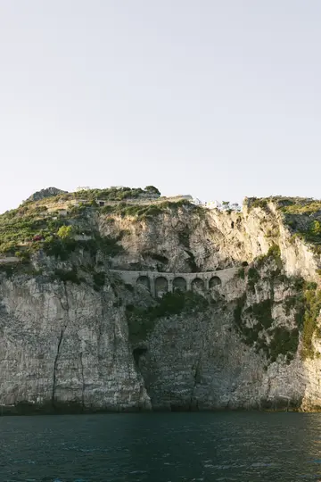 A towering, textured cliff face rises from dark blue water under a pale sky. A stone road with multiple arches is carved…