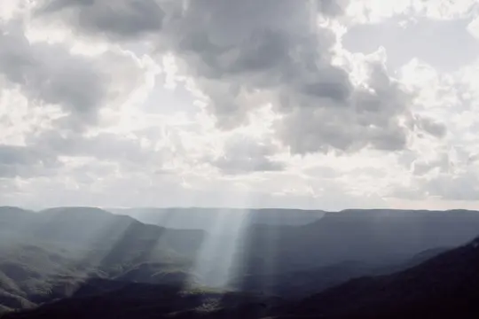 Un'ampia vista panoramica di colline e valli verde scuro coperte di alberi sotto un cielo drammatico con raggi di sole
