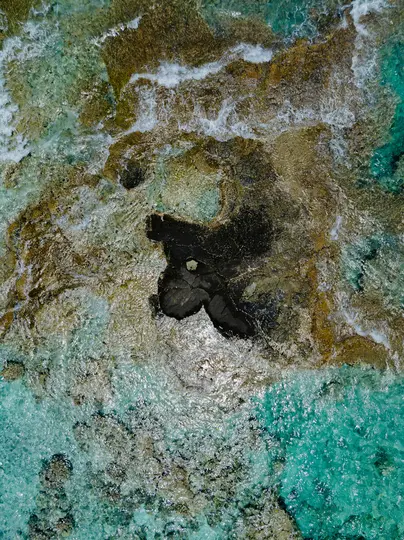 An aerial, top-down view shows clear turquoise ocean water with visible brown and black rock formations and coral reefs.