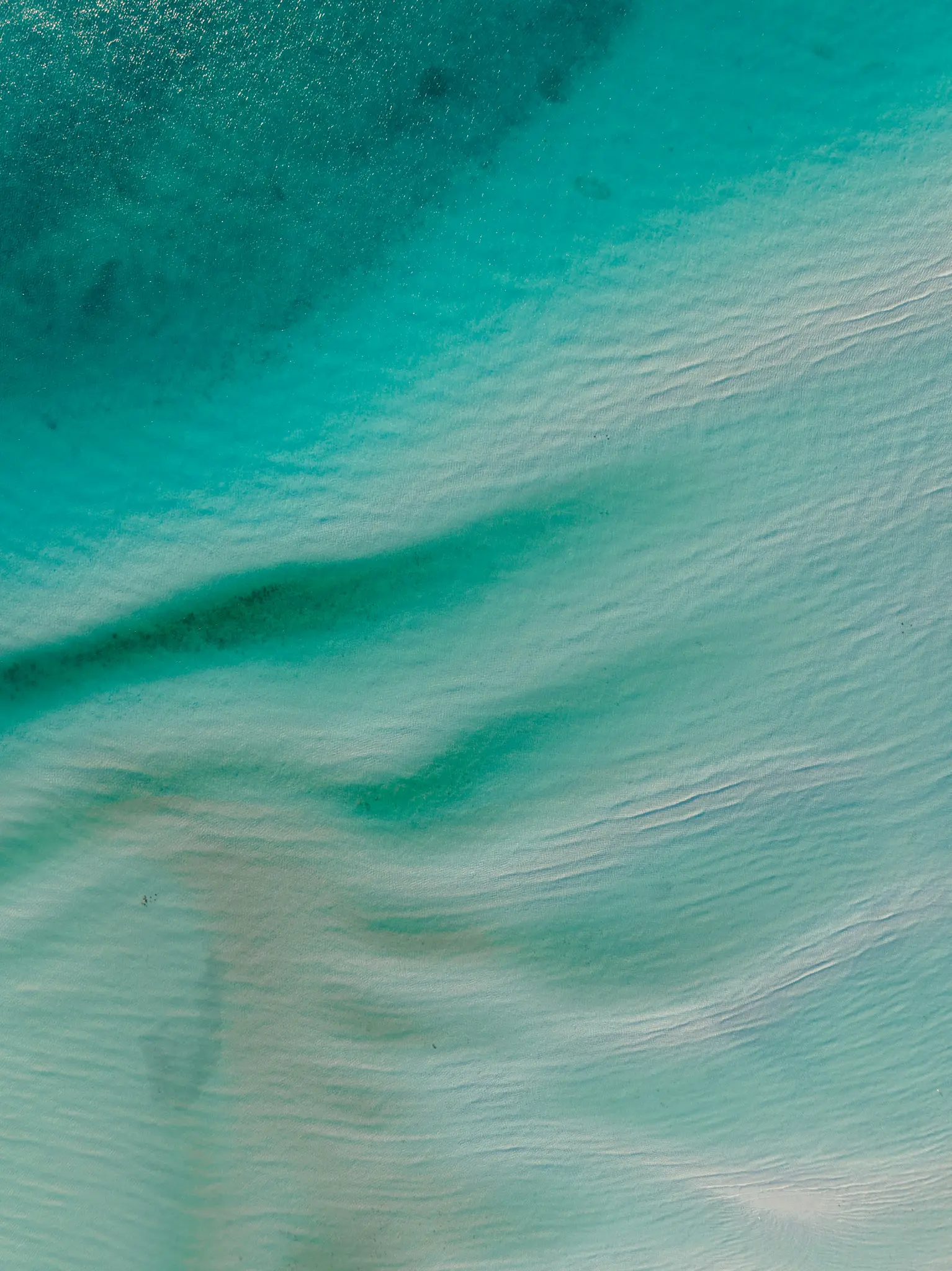 An aerial shot of clear, shallow turquoise ocean water revealing intricate sand ripples on the seabed below.