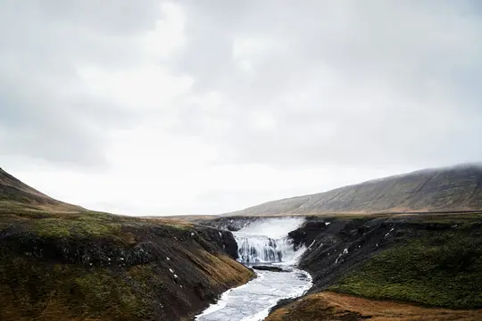 Una ripresa ampia di una cascata che scende in un fiume parzialmente ghiacciato, fiancheggiata da colline muschiose e