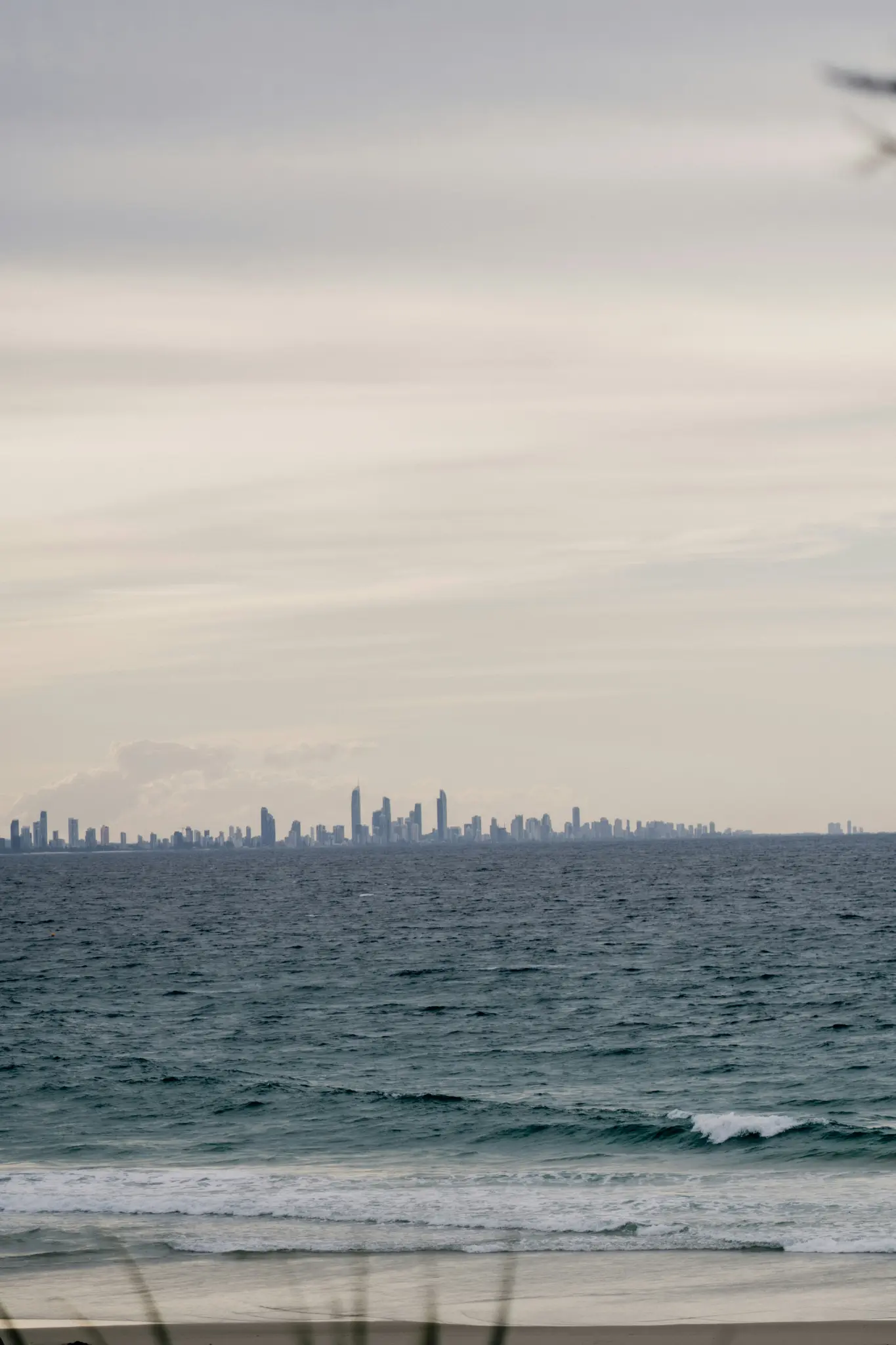 A wide landscape photo showing a distant city skyline with numerous tall buildings across a calm, grey-blue ocean. Gentle…